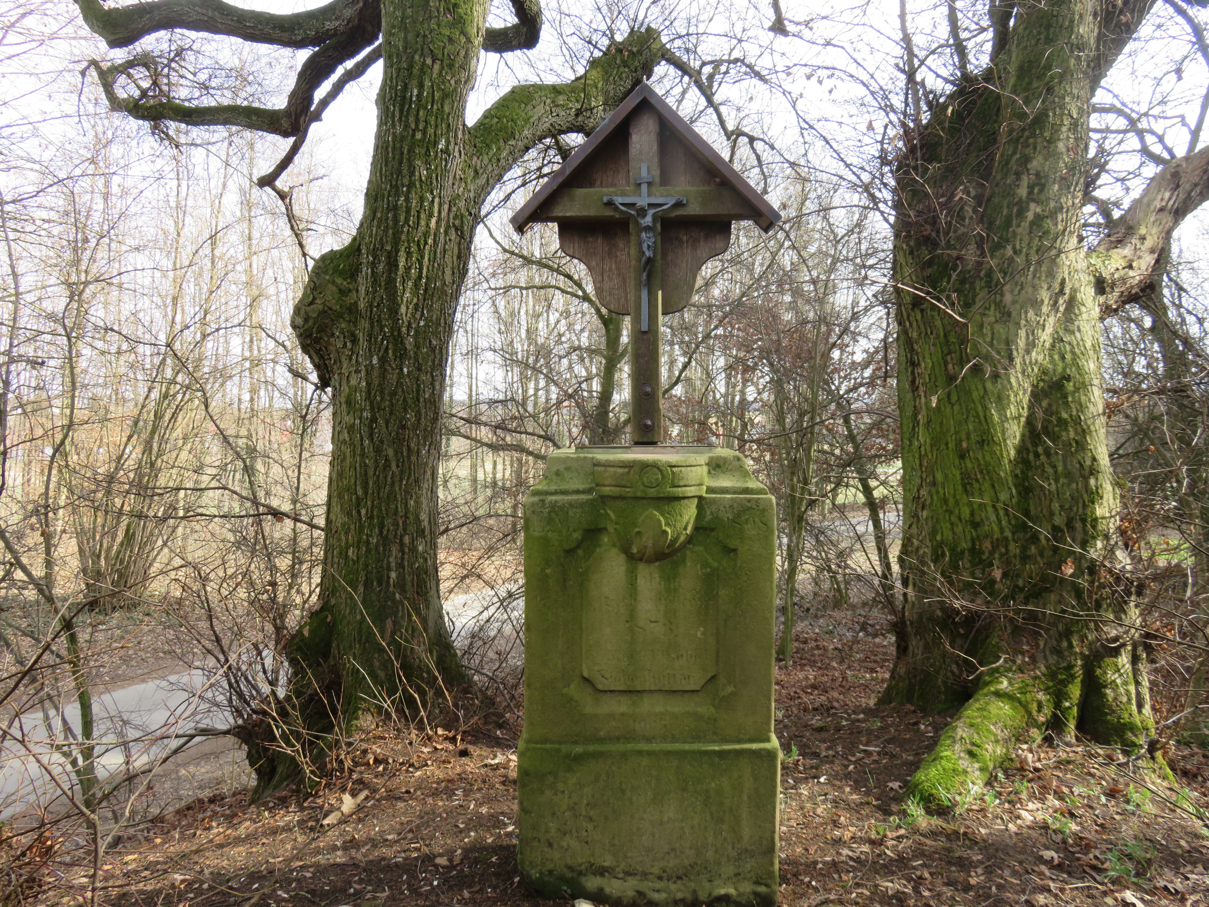 Überdachtes Eichenkreuz mit Dreiecksschild, bronzener Corpus mit Kreuz, Sandsteinsockel. Hattenhofener Holz, an der Gabelung Schmiechener Weg/Prittrichinger Weg.